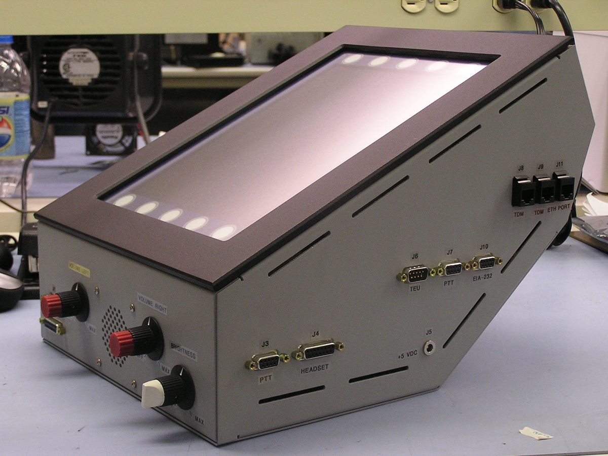 A finished communications console on the test bench with its display backlit and its rear interface connectors labelled.